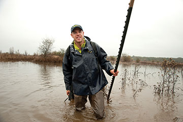 The researcher wading through muddy river water, holding equipment in each hand