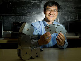 Photo: Cheng holding two modules, sitting in front of blackboard covered with calculations