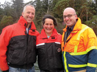 Photo: Two men and a woman, smiling, arm in arm, dressed in rain jackets 