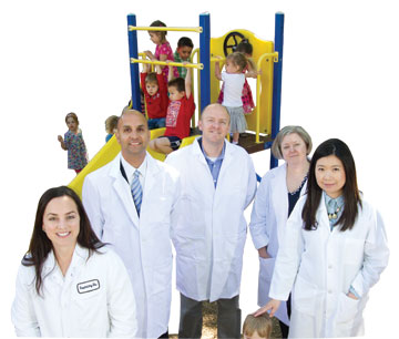 five researchers in lab coats standing, with preschoolers on play structure behind them