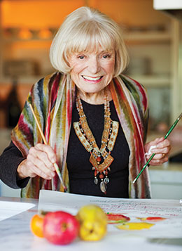 Photo: Margrit Mondavi holding a paintbrush in each hand and standing over a watercolor still life painint of fruit
