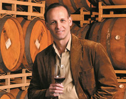Photo: the winemaker holding glass of wine, with barrels in background